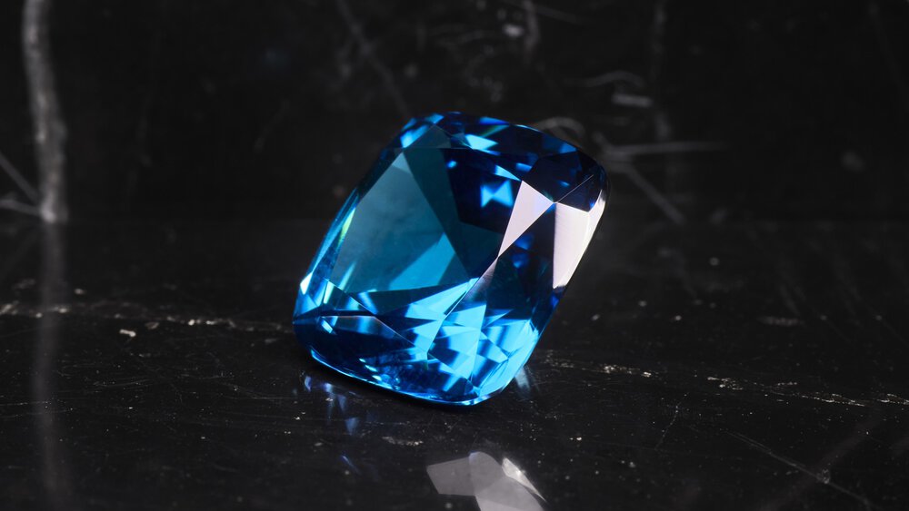 A large, faceted blue gemstone on a reflective black surface.