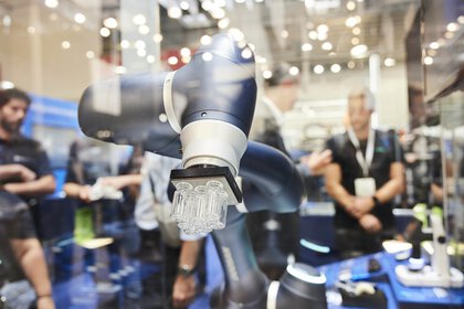 Close-up of a robotic arm on display at an exhibition stand, lifting several small glass bottles with a square surface.