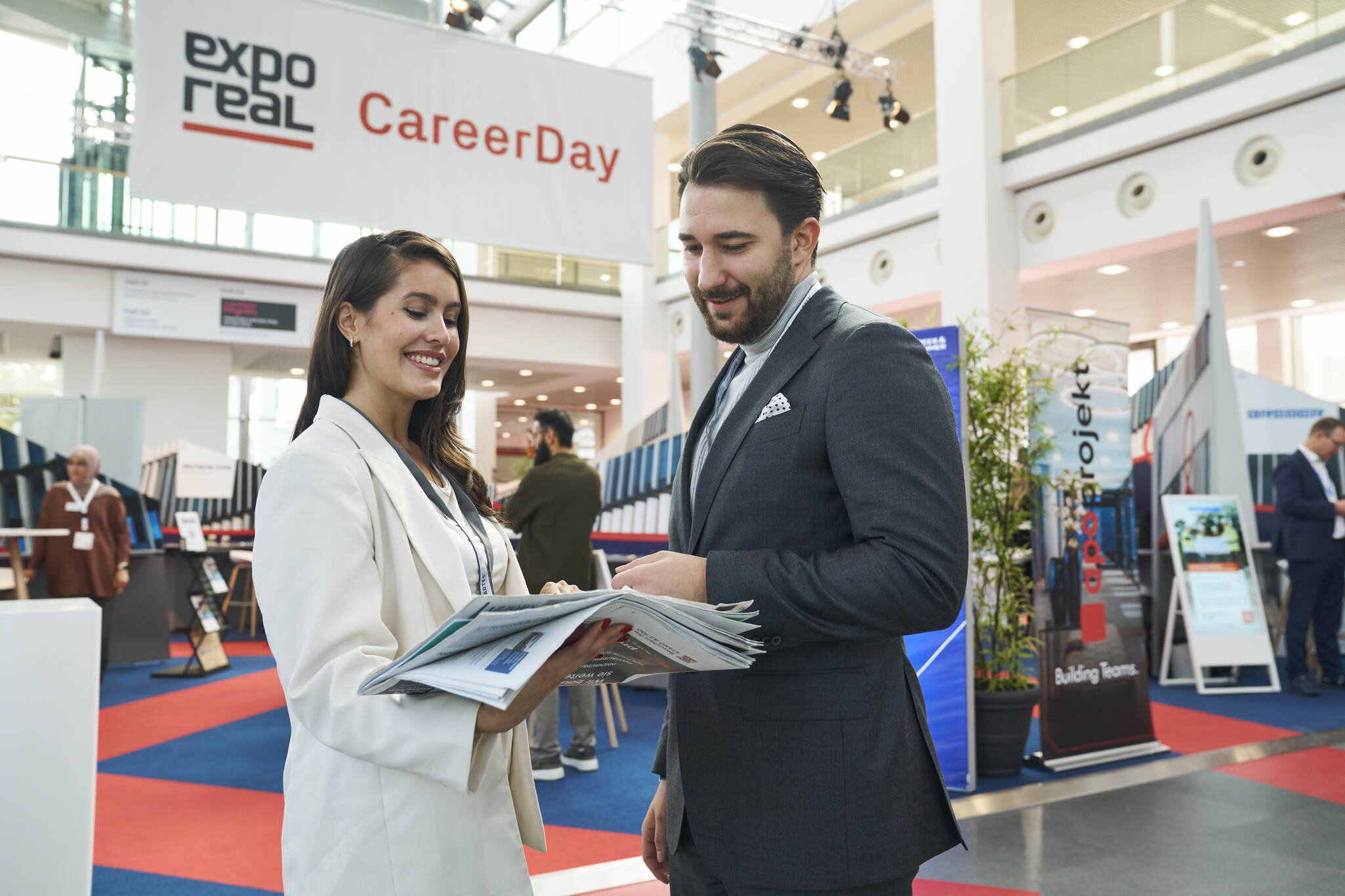 Zwei Personen im Gespräch auf dem „CareerDay“ der EXPO REAL in München. Eine Frau in heller Businesskleidung hält mehrere Broschüren und lächelt, während sie mit einem Mann im Anzug spricht. Im Hintergrund sind Messestände, Infotafeln und ein großes Banner mit der Aufschrift „EXPO REAL CareerDay“ zu sehen.