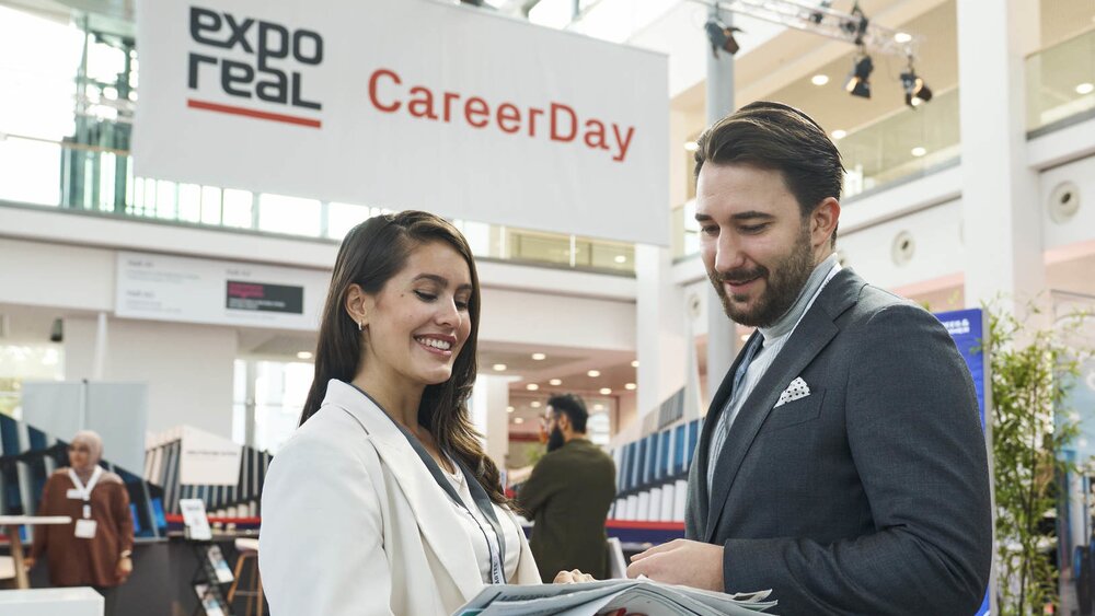 Zwei Personen im Gespräch auf dem „CareerDay“ der EXPO REAL in München. Eine Frau in heller Businesskleidung hält mehrere Broschüren und lächelt, während sie mit einem Mann im Anzug spricht. Im Hintergrund sind Messestände, Infotafeln und ein großes Banner mit der Aufschrift „EXPO REAL CareerDay“ zu sehen.