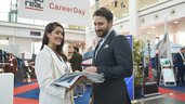 Zwei Personen im Gespräch auf dem „CareerDay“ der EXPO REAL in München. Eine Frau in heller Businesskleidung hält mehrere Broschüren und lächelt, während sie mit einem Mann im Anzug spricht. Im Hintergrund sind Messestände, Infotafeln und ein großes Banner mit der Aufschrift „EXPO REAL CareerDay“ zu sehen.