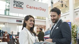 Zwei Personen im Gespräch auf dem „CareerDay“ der EXPO REAL in München. Eine Frau in heller Businesskleidung hält mehrere Broschüren und lächelt, während sie mit einem Mann im Anzug spricht. Im Hintergrund sind Messestände, Infotafeln und ein großes Banner mit der Aufschrift „EXPO REAL CareerDay“ zu sehen.
