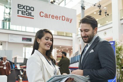 Two people are talking at the “CareerDay” of EXPO REAL in Munich. A woman in light business attire is holding several brochures and smiling while speaking with a man in a suit. In the background, exhibition booths, information displays, and a large banner reading “EXPO REAL CareerDay” can be seen.