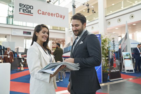 Two people are talking at the “CareerDay” of EXPO REAL in Munich. A woman in light business attire is holding several brochures and smiling while speaking with a man in a suit. In the background, exhibition booths, information displays, and a large banner reading “EXPO REAL CareerDay” can be seen.