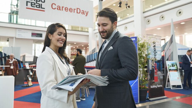 Zwei Personen im Gespräch auf dem „CareerDay“ der EXPO REAL in München. Eine Frau in heller Businesskleidung hält mehrere Broschüren und lächelt, während sie mit einem Mann im Anzug spricht. Im Hintergrund sind Messestände, Infotafeln und ein großes Banner mit der Aufschrift „EXPO REAL CareerDay“ zu sehen.