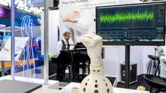 A mechanical hand with a white glove in front of a screen with data visualizations. A person at a piano can be seen blurred in the background.
