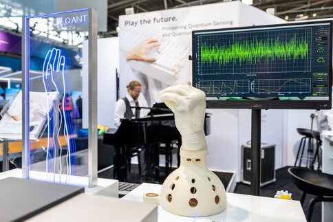 Eine mechanische Hand mit weißem Handschuh vor einem Bildschirm mit Datenvisualisierungen. Im Hintergrund ist unscharf eine Person an einem Klavier zu sehen.