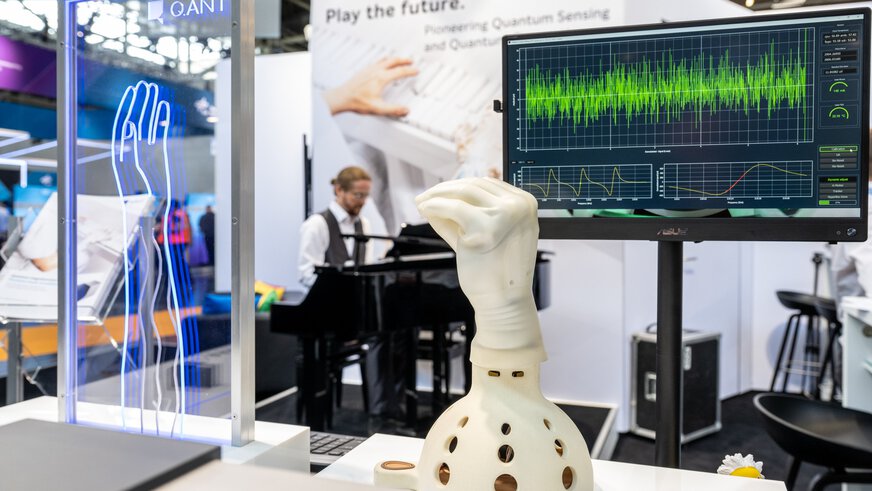 A mechanical hand with a white glove in front of a screen with data visualizations. A person at a piano can be seen blurred in the background.