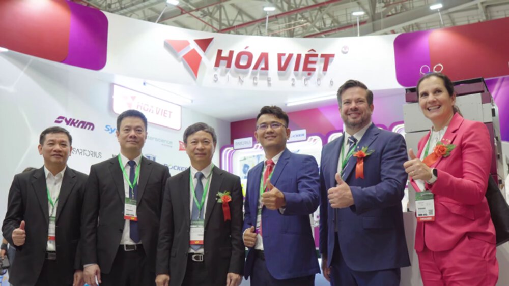 Six people in suits are standing in front of a Hóa Việt stand, smiling and giving a thumbs-up – it appears to be a trade fair or exhibition.