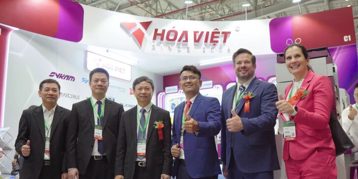 Six people in suits are standing in front of a Hóa Việt stand, smiling and giving a thumbs-up – it appears to be a trade fair or exhibition.