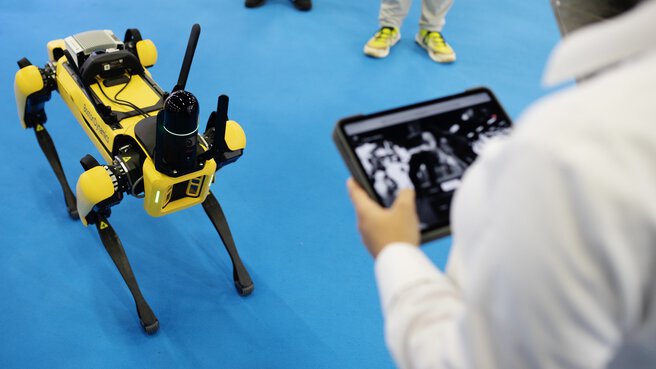 Ein gelber Roboterhund steht vor einer Person mit einem Tablet, auf dem ein Schwarzweißbild aus der Kamera des Roboters zu sehen ist.