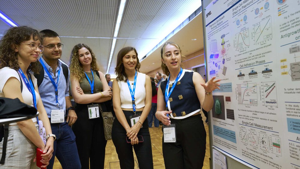 A group of people listen attentively to a woman presenting a scientific poster.