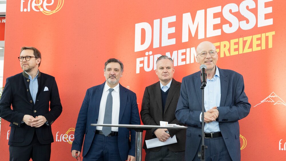 Four men in suits stand in front of an orange trade fair backdrop with the words “The trade fair for my leisure time” during an official opening ceremony, while one of the men speaks into a microphone.