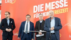 Four men in suits stand in front of an orange trade fair backdrop with the words “The trade fair for my leisure time” during an official opening ceremony, while one of the men speaks into a microphone.