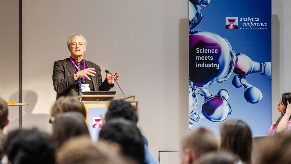 A man with grey hair and a striped jacket gives a presentation to an audience at the analytica conference.