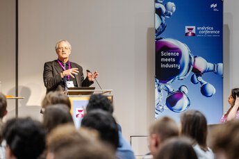 A man with grey hair and a striped jacket gives a presentation to an audience at the analytica conference.