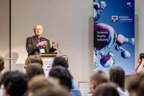 Ein Mann mit grauen Haaren und im gestreiften Sakko hält auf der analytica Conference einen Vortrag vor Publikum.