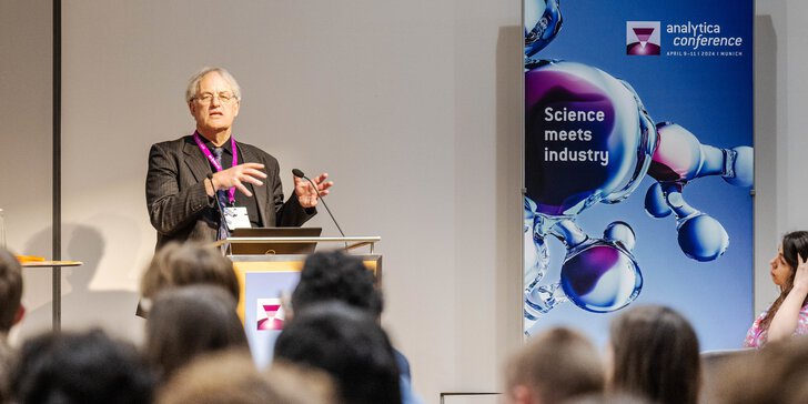 A man with grey hair and a striped jacket gives a presentation to an audience at the analytica conference.