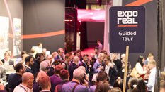 A large group of visitors participates in a guided tour at EXPO REAL in Munich. In the foreground, a sign reads “expo real – Guided Tour.” Some participants are wearing headphones and listening to explanations as they move through the trade fair. In the background, more exhibition booths and people engaged in conversations can be seen.