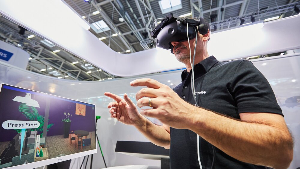 A man at a trade fair stand wears VR glasses and stands gesticulating next to a screen that shows his point of view.