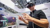 Ein Mann an einem Messestand trägt eine VR-Brille und steht gestikulierend neben einem Bildschirm, der seinen Blickwinkel zeigt.