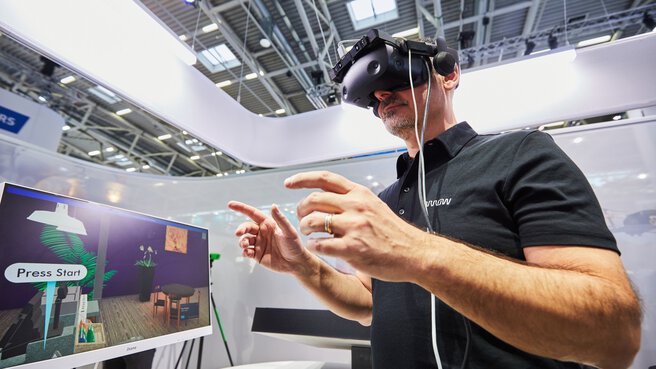 Ein Mann an einem Messestand trägt eine VR-Brille und steht gestikulierend neben einem Bildschirm, der seinen Blickwinkel zeigt.