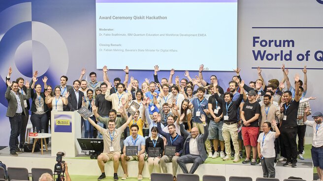 Gruppenfoto bei der Award Ceremony des Qiskit Hackathons auf der World of Quantum 2025 in München, alle jubeln auf der Bühne.