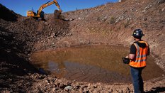 A worker in protective clothing watches an excavator working near a partially water-filled pit.
