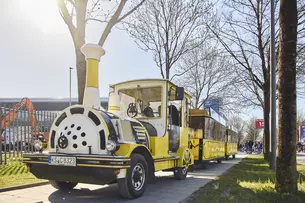 Ein gelber Zug im Eisenbahnlook fährt auf dem Weg neben einer Wiese mit Bäumen.