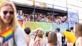 A group of people on a colourful parade float are waving to spectators in summery, rainbow-coloured outfits.