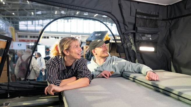 A man and a woman are inquiring about a camping or roof tent module at a trade fair booth and examining the interior features.