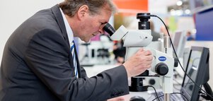 Ein Mann im Anzug sieht an einem Messestand durch ein Mikroskop.