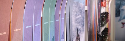 Bunte Skier stehen vertikal aufgereiht an einer Ausstellerwand, im Hintergrund sind Skiausrüstung und Bergfotos zu sehen.
