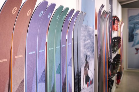 Bunte Skier stehen vertikal aufgereiht an einer Ausstellerwand, im Hintergrund sind Skiausrüstung und Bergfotos zu sehen.