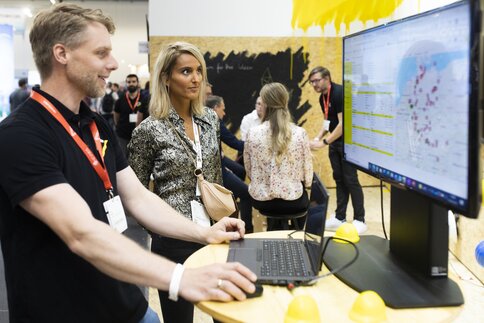 Zwei Personen stehen an einem Tisch und schauen auf einen Computerbildschirm eine Landkarte mit Markierungen an, weitere Personen sitzen im Hintergrund.