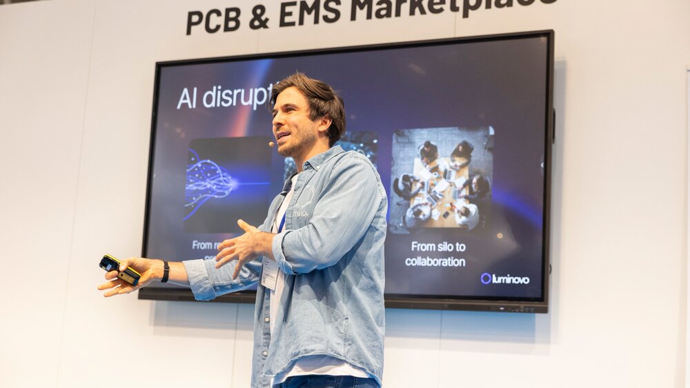 Ein Mann mit braunen Haaren in Jeansbekleidung hält ein kleines Gerät in der Hand und hält vor einem Bildschirm einen Vortrag.  Über ihm steht „PCB & EMS Marketplace“.