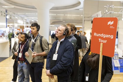 A group of people wearing headphones are taking part in a guided tour at a trade fair; a sign reads “Rundgang Tour” (guided tour).