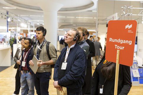 A group of people wearing headphones are taking part in a guided tour at a trade fair; a sign reads “Rundgang Tour” (guided tour).