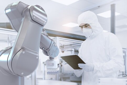 A scientist in protective clothing uses a tablet near a robotic arm in a modern laboratory