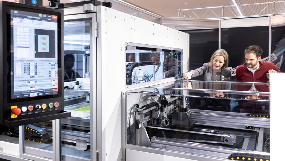A man and a woman look inside a large transparent machine with lots of cables, belts and a control panel with numerous buttons.