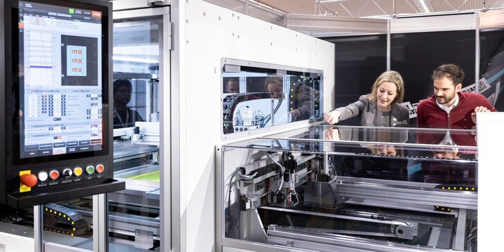 A man and a woman look inside a large transparent machine with lots of cables, belts and a control panel with numerous buttons.