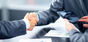Two people in suits shake hands over a table with documents.