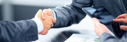 Two people in suits shake hands over a table with documents.
