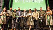 Nine people in festive attire stand in front of the green INTERFORST logo and cut the green and white opening ribbon together.