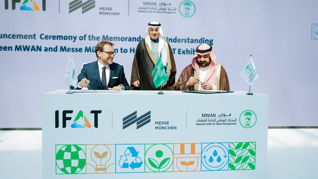 Zwei Männer unterschreiben Dokumente an einem Tisch mit IFAT- und MWAN-Logos; ein weiterer Mann steht lächelnd hinter ihnen.