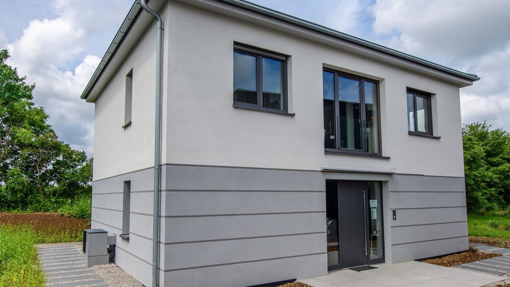 Modernes zweistöckiges Haus mit Flachdach, grau-weißer Fassade, großen Fenstern und gepflastertem Weg.