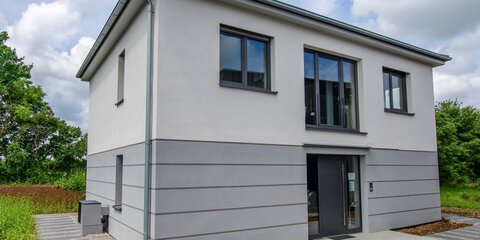 Modernes zweistöckiges Haus mit Flachdach, grau-weißer Fassade, großen Fenstern und gepflastertem Weg.