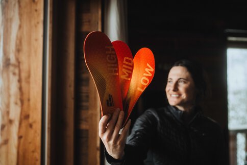 Eine Frau in einer schwarzen Jacke hält lächelnd drei orangene Einlegesohlen in der Hand, auf denen jeweils die Worte „HIGH“, „MID“ und „LOW“ stehen.