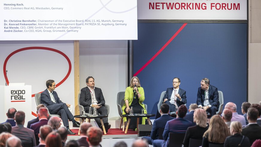 A panel discussion at the “Discussion & Networking Forum” during EXPO REAL in Munich. Four experts are seated on stage discussing the German real estate and investment market in 2030, while the audience listens attentively. A female speaker in a green jacket holds a microphone and speaks, with the other panelists seated beside her. Behind them, a screen displays the discussion topic and the participants’ names.