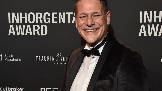 A man in a tuxedo holds the trophy for the “Luxury Watch of the Year” category and smiles in front of a logo wall at the AWARD Gala.